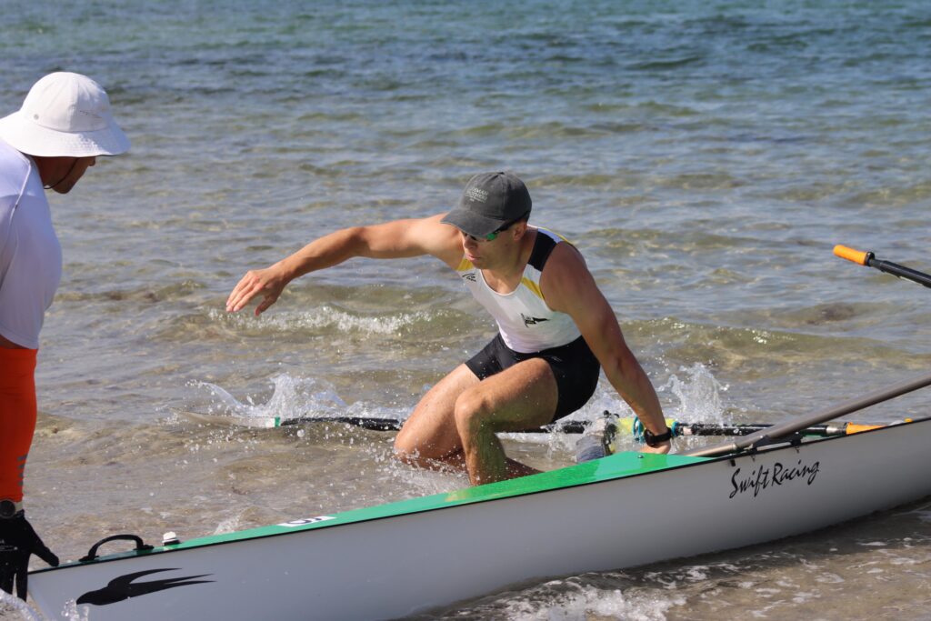 Zwei Personen am Ufer; eine steigt aus einem sportlichen Ruderboot mit der Aufschrift "Swift Racing".